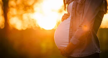 Pregnant woman cradling belly outdoors, bathed in warm sunset light.