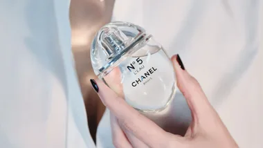 A hand with black nails holding a bottle of Chanel No. 5 L'Eau perfume against a white background.