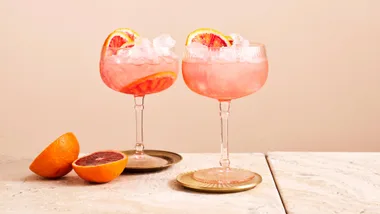 Two pink cocktails in glasses with blood orange slices, on a marble surface, next to halved blood oranges.