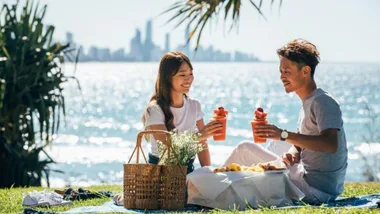 A couple enjoys a picnic by the beach with tropical drinks, against a city skyline in the background.