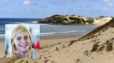 Blonde woman smiling in foreground, sandy beach and ocean with green hills in background.