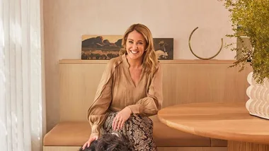 A woman smiling at a table with a dog beside her, in a warmly lit room with art in the background.