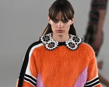 Model with long brown hair wearing an orange fuzzy sweater and black skirt on the runway.