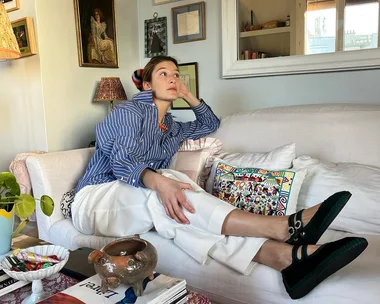 A person in a striped shirt and white pants sits on a sofa, surrounded by framed art on the wall and a coffee table with magazines.