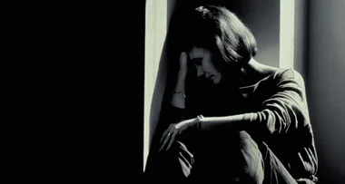 A woman sits in a dark corner, resting her head on her hand, seemingly overwhelmed and distressed.