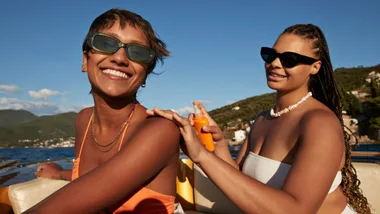Two people on a boat, one applying sunscreen to the other's back, both wearing sunglasses and smiling.