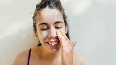 Person smiling and applying cream to their face with eyes closed against a light background.