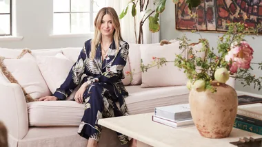 A woman in floral pajamas sits on a sofa in a cozy living room with a vase of flowers and books on the table.