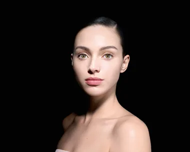 Portrait of a woman with slicked-back hair against a black background, showcasing clear skin and neutral expression.